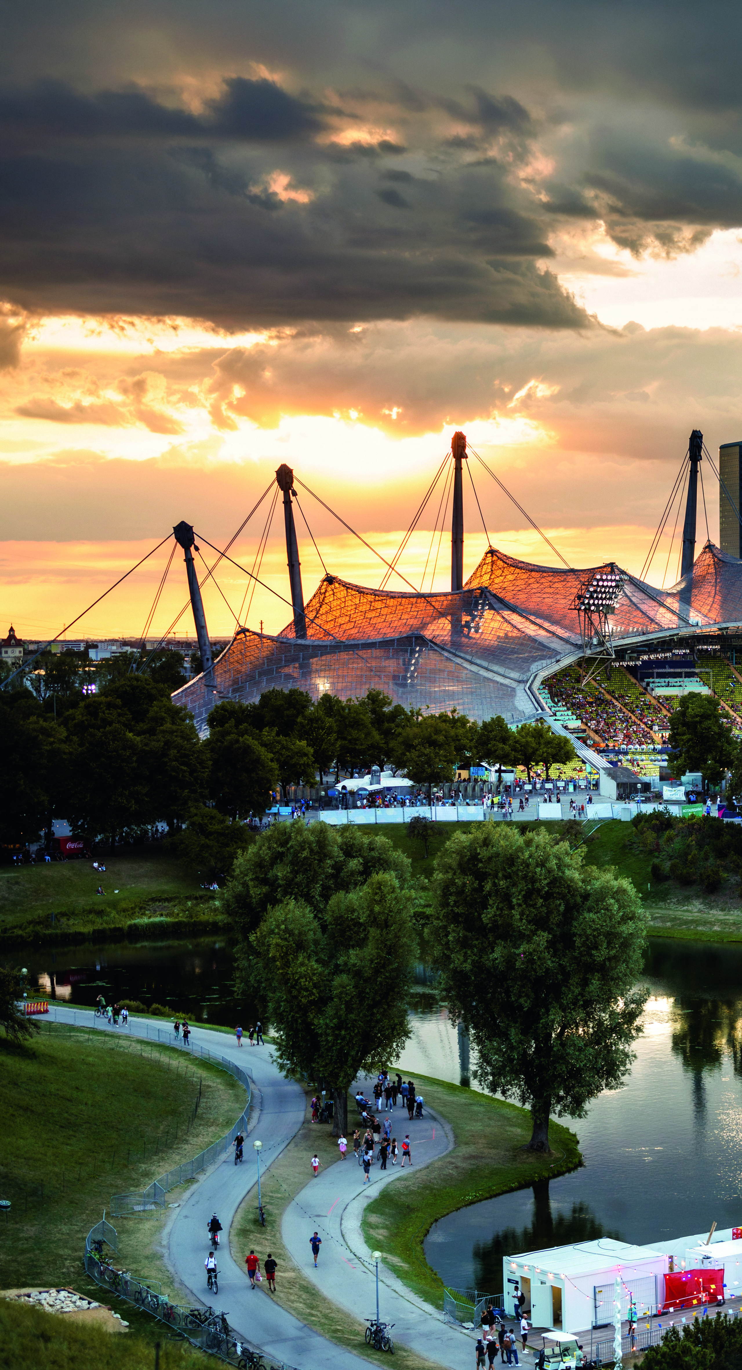 Sonnenuntergang über dem Olympiastadion in München mit See und entspannten Passanten.
