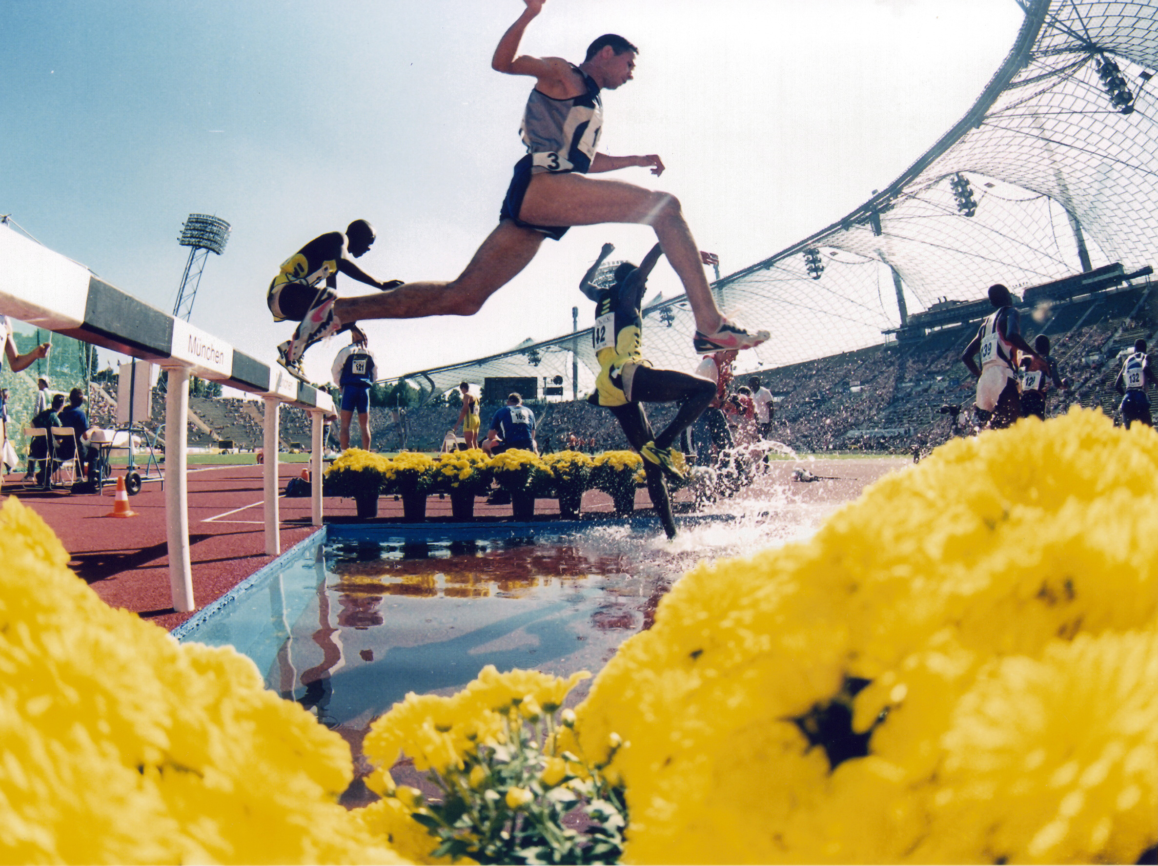 Hürdenlauf Wettbewerb im Rahmen der Olympischen Spiele 1972 in München. Perspektive von unten aufgenommen. Zwei Athleten springen über eine Hürde und Wassergraben. Im Hintergrund sieht man die volle Tribüne und das Zeltdach des Olympiastadions